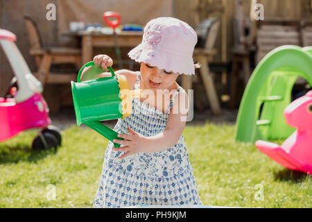 Vorderansicht eines kleinen Mädchens, der in der Rückseite Garten mit lässige Kleidung und ein Sonnenhut auf. Sie spielt mit einem Spielzeug Gießkanne und t Stockfoto