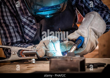 Nahaufnahme eines jungen Mannes in Uniform, schweißen Schweißer-Maske und Schweißer Leder, Schweißgut mit einem Arc Welding Machine in Werkstatt, Blau und Orange Stockfoto