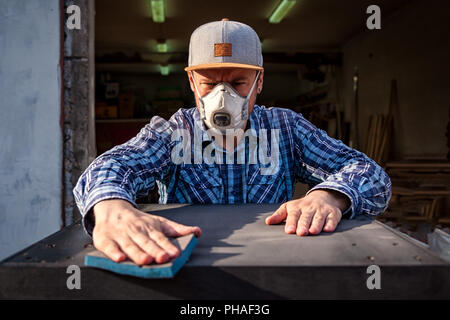 Starke Mann in Arbeitskleidung bemüht und wäscht, Polituren einen Lappen mit einem hölzernen schwarz Tisch in der Werkstatt Stockfoto