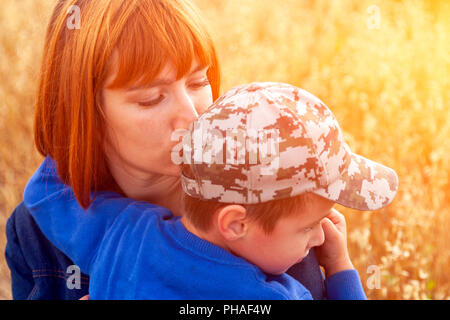 Schöne Frau und ihr kleiner Sohn zittern und stark annehmen, im Feld im Hintergrund. Sommer Portrait von Mutter und Sohn am Muttertag. Stockfoto