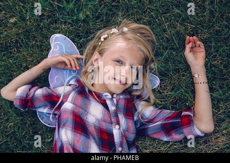 Portrait von lächelnden blonde weiße Kaukasier Kind Mädchen mit langem Haar zu tragen rosa Fee Flügel und Plaid Shirt, liegend auf Gras im Park, Ansicht von t Stockfoto