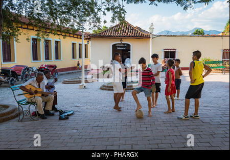 Trinidad, Kuba/März 15, 2016: Junge Jungen Fußball spielen in einer Stadt wie zwei Männer, die traditionelle Kubanische Musik auf Gitarre spielen. Stockfoto