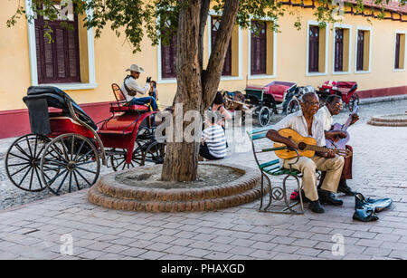 Trinidad, Kuba/März 15, 2016: Zwei kubanischen Gitarristen auf einer Bank spielen Gitarren an einem Platz in der UNESCO-Stadt Trinidad sitzen; eine Reihe von Pferd und Bu Stockfoto