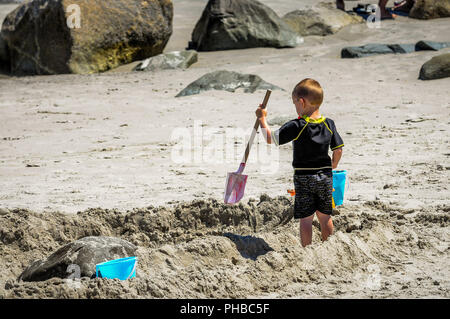 Dieser kleine Junge hält seinen Eimer in seiner rechten Hand, und mit seiner linken Er gräbt sie dieses Big Rock. Jetzt übergab er einen Graben auf den Ozean zu graben Stockfoto