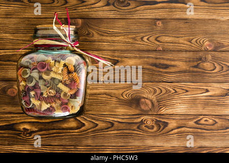 Klares Glas storage Jar mit Makkaroni fallen Pasta in einer Mischung aus Formen und Farben auf Ihrer Seite auf einen hölzernen Hintergrund mit Kopie Raum Stockfoto