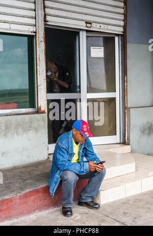 Havanna, Kuba/März 21, 2016: Trotz Cubas begrenzte WLAN-Konnektivität, um einen Mann-über-tragen eines Kuba Baseball Hut - sitzt auf einer Tür mit einem Smartphone. Stockfoto