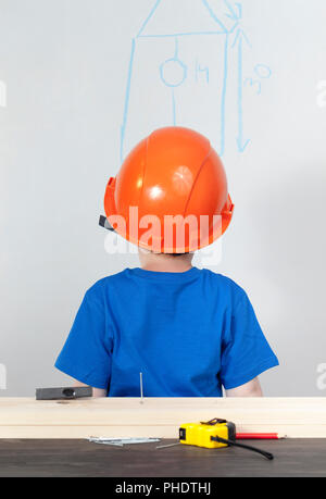 Ein Junge in einem orange Helm und einem blauen T-Shirt aufpaßt, wie es notwendig ist, ein Vogelhaus, hintere Ansicht von der Rückseite zu errichten, Stockfoto