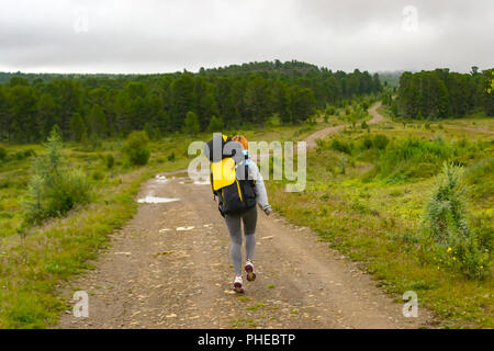 Mädchen reisen mit großen schwarzen Rucksack und eine Flasche Wasser in sie gehen auf der Straße, entlang des Waldes mit grünen Nadelbäume zu den Bergen und zu den Advent Stockfoto