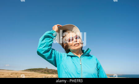 Attraktive reife asiatische Dame trägt blaue Anorak, canvas Hut, Sonnenbrille. Sieht bis zu blauen Himmel mit Mimik für Interpretation. Stockfoto