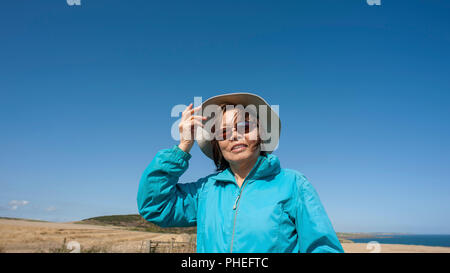 Attraktive reife asiatische Dame trägt blaue Anorak, canvas Hut, Sonnenbrille. Sieht bis zu blauen Himmel mit Mimik für Interpretation. Stockfoto