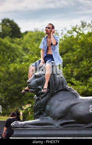 Junger Mann sitzt auf der Spitze eines Lion Skulptur und hört Musik mit seinen Kopfhörern in Mount Royal Park, Montreal, Kanada. Stockfoto