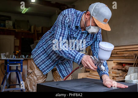 Erfahrene Tischler in Arbeitskleidung und kleine Steuerungsfunktionen für Eigentümer malt eine Holzkiste aus der Kommode in der Farbe schwarz in der Werkstatt, im Hintergrund eine Menge Stockfoto