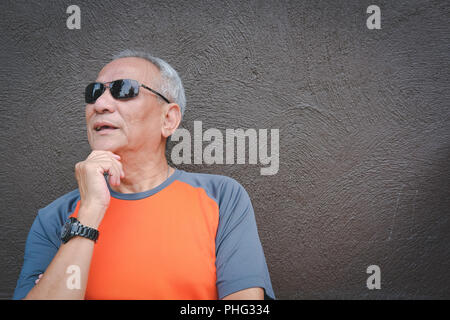 Nachdenklich ältere Mann mit Sonnenbrille in der Nähe von Brown Wand. Die asiatischen ältere männliche Denken. senior Freizeitaktivitäten lifestyle Stockfoto