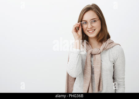 Taille-up Shot von attraktiven stilvolle junge Frau in Gläsern und rosa Pullover über Hals tragen Bluse berühren Rahmen und lächelt freundlich in die Kamera hanging out in unbekannten Firma Stockfoto