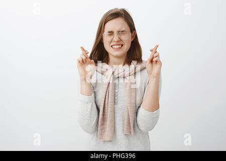 Taille-up Shot der nervösen good-looking Smart Girl in Bluse und Pullover über Hals schließen Augen gebunden und Aufeinanderpressen der Zähne während Gefühl intensiven drückt die Daumen für gutes Glück zu wünschen über graue Wand Stockfoto