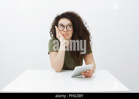 Portrait von Verträumten sorglos schöne Studentin in schwarz Brille, den Blick nach oben und schiefen Gesicht auf Hand, während Sie am Tisch sitzen, halten digitale Smartphone, Langeweile und gleichgültig Stockfoto