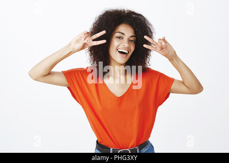 Fertige positive Emotionen von heute zu entdecken. Gerne sorglos attraktive Frau mit dunkler Haut und Afro Frisur, breit und lächelnd Sieg oder Frieden Geste in der Nähe von Gesicht, über graue Wand stehend Stockfoto