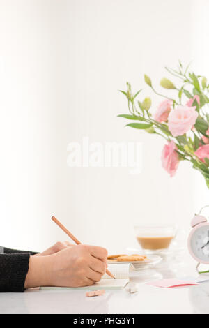 Weibliche Hand schreiben in einem Notebook auf dem Schreibtisch, Ansicht von oben. Auf dem weißen Tisch legen Blumen Stockfoto