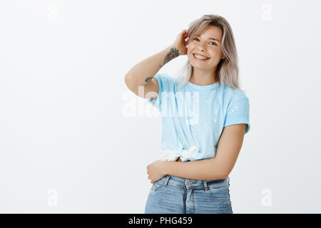 Portrait von attraktiven fröhliche Blondine mit Tattoos im stilvollen Outfit, berühren Haar und breit grinsend, Erröten oder erfreut sein, Kompliment, tolle Stimmung während über graue Wand posieren Stockfoto