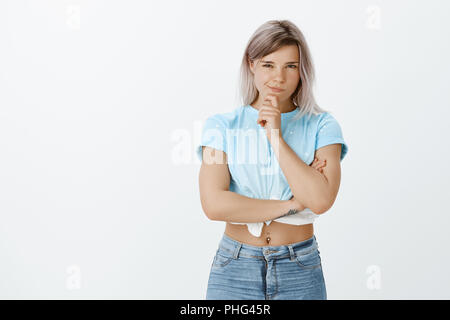 Ich habe Pläne für Sie. Portrait von fasziniert nachdenklich charmante Frau mit blonden Haaren und durchbohrte Bauch, Schielen und schürzte die Lippen während Hand am Kinn, Denken oder in Idee über graue Wand Stockfoto