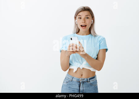 Es heraus überprüfen, Klang ist erstaunlich. Beeindruckt und erfreut, attraktive Frau mit blondem Haar in der stilvollen Kleidung, den Blick mit Erstaunen an der Kamera während der Verwendung von neuen Smartphones und drahtlose Kopfhörer Stockfoto