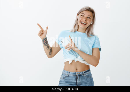 Charmante positive weibliche Kursteilnehmer mit blondem Haar und tätowiert arm, hören Musik in drahtlosen Ohrhörer und Holding smartphone, zeigen und sich an der oberen, linken Ecke über graue Wand Stockfoto
