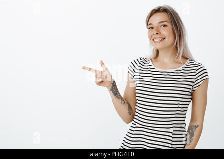 Portrait von Happy charmante Frau mit blondem Haar und Tätowierungen, lächelte breit und zeigt mit dem Finger gun Geste links, Anzeigen toller Ort zum Übernachten und Entspannen. Über grauer Hintergrund posiert Stockfoto