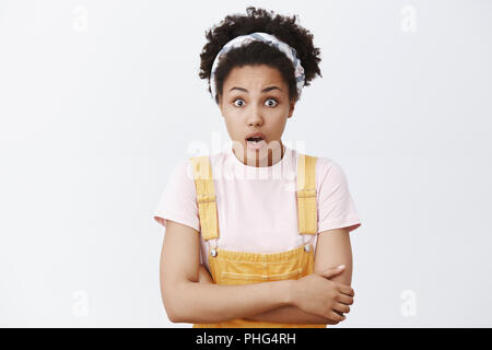 Mitarbeiter sprechen über neue Gerücht verbreiten über Büro, schockiert. Schüttelte beeindruckt African American Girl in Kopfbügel und gelbe Overalls, halten die Hände auf der Brust und mit Blick auf die Kamera Stockfoto