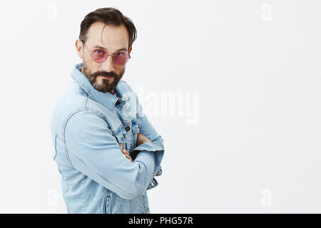 Innen- schuss von Ernst - auf der Suche lustige und gefühlvolle stilvolle homosexuellen Mann in Jeans Jacke, Sonnenbrille, ständigen halb umgedreht grauer Hintergrund in zuversichtlich Pose mit der Hand auf der Brust gekreuzt, hinting Stockfoto