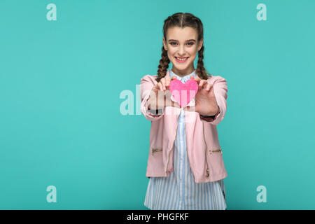 Happy schönes Mädchen in lässigen Stil, pigtail Frisur und rosa Jacke, stehend und mit rosa Herz und Kamera mit toothy Lächeln Stockfoto