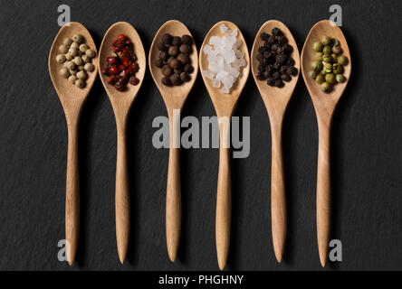 Verschiedene Arten von Pfeffer und Salz in Holzlöffel auf schwarzem Schiefer Hintergrund Stockfoto