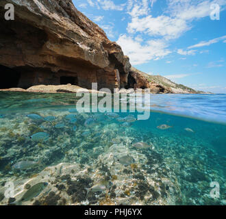 Felsige Küste mit Höhle am Meer und Schwarm von Fischen unter Wasser, geteilte Ansicht oberhalb und unterhalb der Wasseroberfläche, Mittelmeer, Costa Blanca, Javea, Spanien Stockfoto