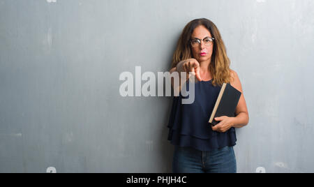 Mittleres Alter Hispanic Frau stehend über Grau grunge Mauer, die ein Buch mit dem Finger an die Kamera und an Sie gerichtet, Handzeichen, positive und Vertrauen Stockfoto