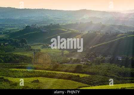 Blick auf die Hügel der Langhe und Schloss von Grinzane Cavour bei Sonnenuntergang, der Himmel ist gelb und orange und einige Sonnenstrahlen den Landsitz in der Hintergrundbeleuchtung aufleuchten Stockfoto