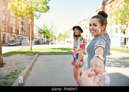 Zwei attraktive junge Freundinnen lächelnd und Führung einer Person durch die Hand beim Bummel durch die Stadt an einem sonnigen Tag Stockfoto