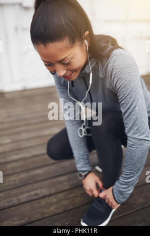 Lächelnde junge asiatische Frau in Sportkleidung kniend auf dem Boden außerhalb ihrer Schnürsenkel binden, bevor sie für einen Lauf Stockfoto