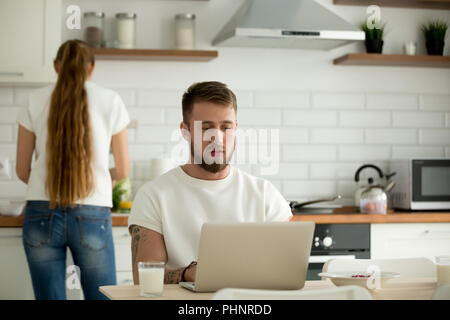 Mann mit Laptop, während Frau Frühstück zubereiten in der Küche Stockfoto