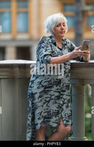 Trendy reife Frau lehnte sich auf eine steinerne Brüstung mit ihrem Mobiltelefon eine SMS an einen Freund zu senden Stockfoto