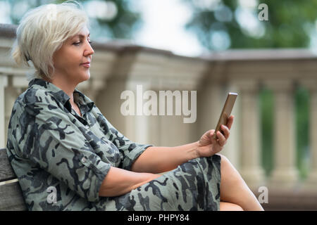 Attraktive Frau sitzt auf einer Bank im Freien lesen auf Ihrem Mobiltelefon mit einem ruhigen Lächeln der Freude in einer Nahaufnahme, Seitenansicht Stockfoto