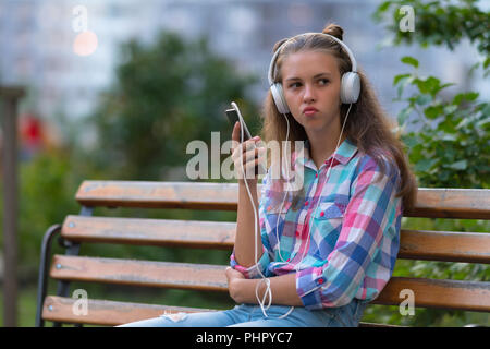 Nachdenkliche junge Frau ihre Lippen schürzte, als sie auf einer Parkbank sitzt, hören die Musik auf Stereo-kopfhörer mit Ihrem Handy Stockfoto