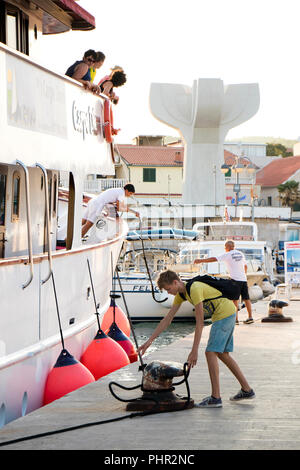 Vodice, Kroatien - 11. August 2018: ein Kreuzfahrtschiff Carpe Diem Ankunft in Vodice und Andocken. Ein Mann und ein Teenager auf einem Pier Befestigung das Schiff Seil Stockfoto