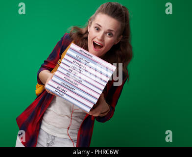 Gerne moderne student Frau im roten Hemd mit Rucksack und Kopfhörer balancing einen Stapel Bücher auf grünem Hintergrund isoliert Stockfoto