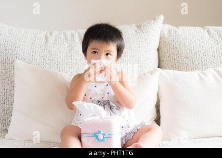 Kranken kleinen asiatischen Mädchen wischen oder Reinigung der Nase mit einem Taschentuch, auf dem Sofa zu Hause sitzen. Medizin und Gesundheit Konzept. Stockfoto