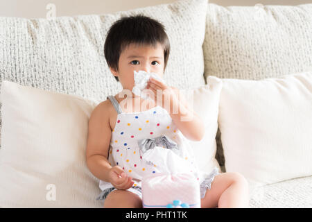 Kranken kleinen asiatischen Mädchen wischen oder Reinigung der Nase mit einem Taschentuch, auf dem Sofa zu Hause sitzen. Medizin und Gesundheit Konzept. Stockfoto