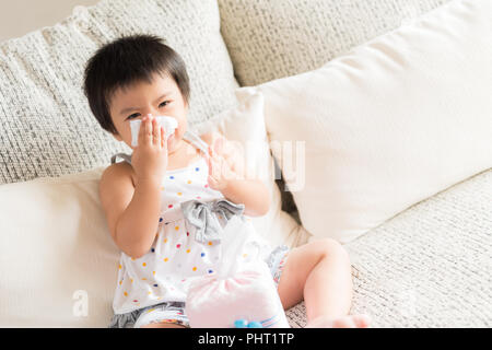 Kranken kleinen asiatischen Mädchen wischen oder Reinigung der Nase mit einem Taschentuch, auf dem Sofa zu Hause sitzen. Medizin und Gesundheit Konzept. Stockfoto