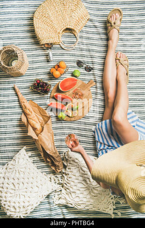 Sommer Picknick Einstellung. Junge Frau im gestreiften Kleid und Stroh Sonnenhut sitzend mit einem Glas Wein in der Hand Rose, frisches Obst an Bord und Baguette auf bla Stockfoto