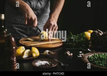 Ein Mann Schichten eine Zitrone und bereitet das Mittagessen mit Artischocken und Erbsen Stockfoto