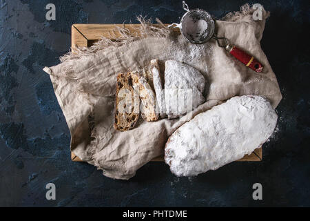 Ganze und aufgeschnittene Weihnachten Kuchen, traditionelle deutsche Festliches backen. Vollkorn Stollen mit Rosinen und Zucker Pulver auf Leinen Servietten, Holz- fach, si Stockfoto