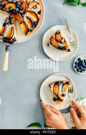 Einer Frau mit einem Schnitt von Pfirsich Kuchen mit Blaubeeren auf einem weißen Keramikplatte und eine Gabel auf einem grauen Hintergrund von der Ansicht von oben fotografiert. Ein weiterer sl Stockfoto