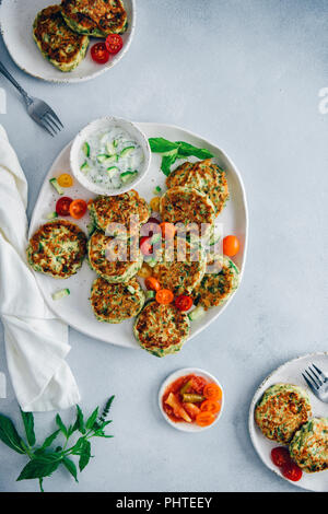 Zucchini Krapfen auf einer Keramikplatte mit Trauben Tomaten und Joghurt dip auf der Seite von der Ansicht von oben fotografiert. Zwei kleinere Platten und Frische mi Stockfoto